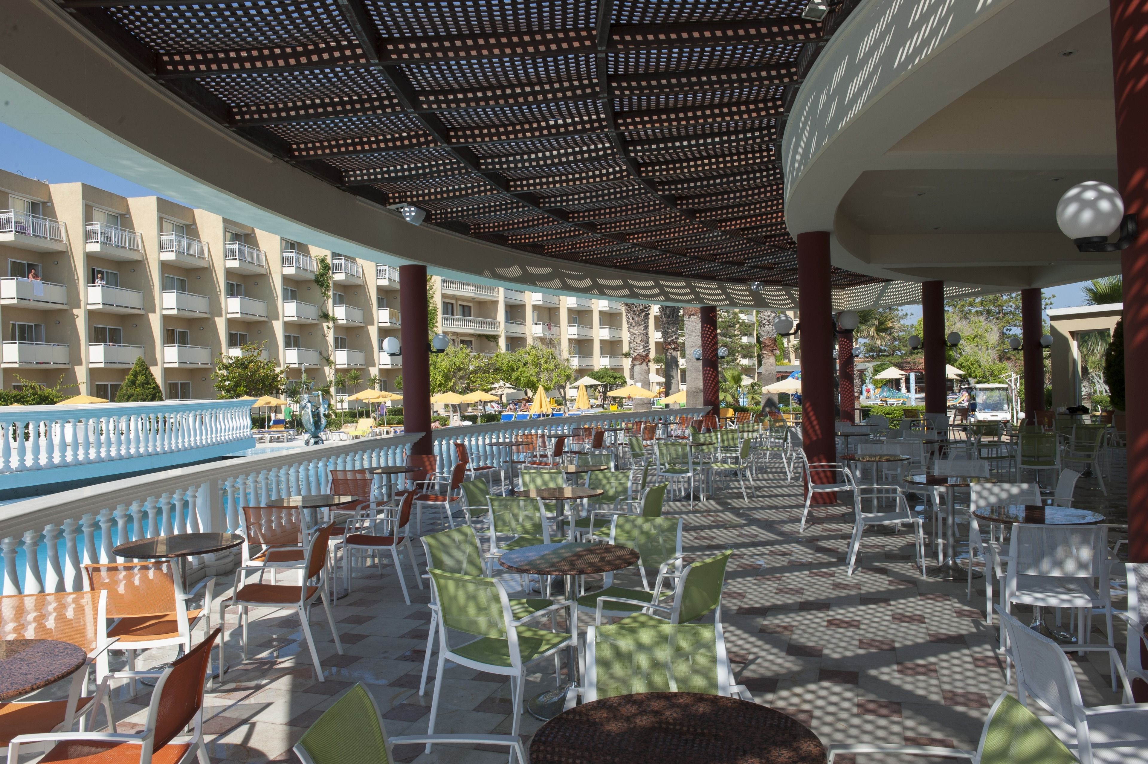 An outdoor cafe area with tables and chairs, a pool, and a building with balconies in the background.