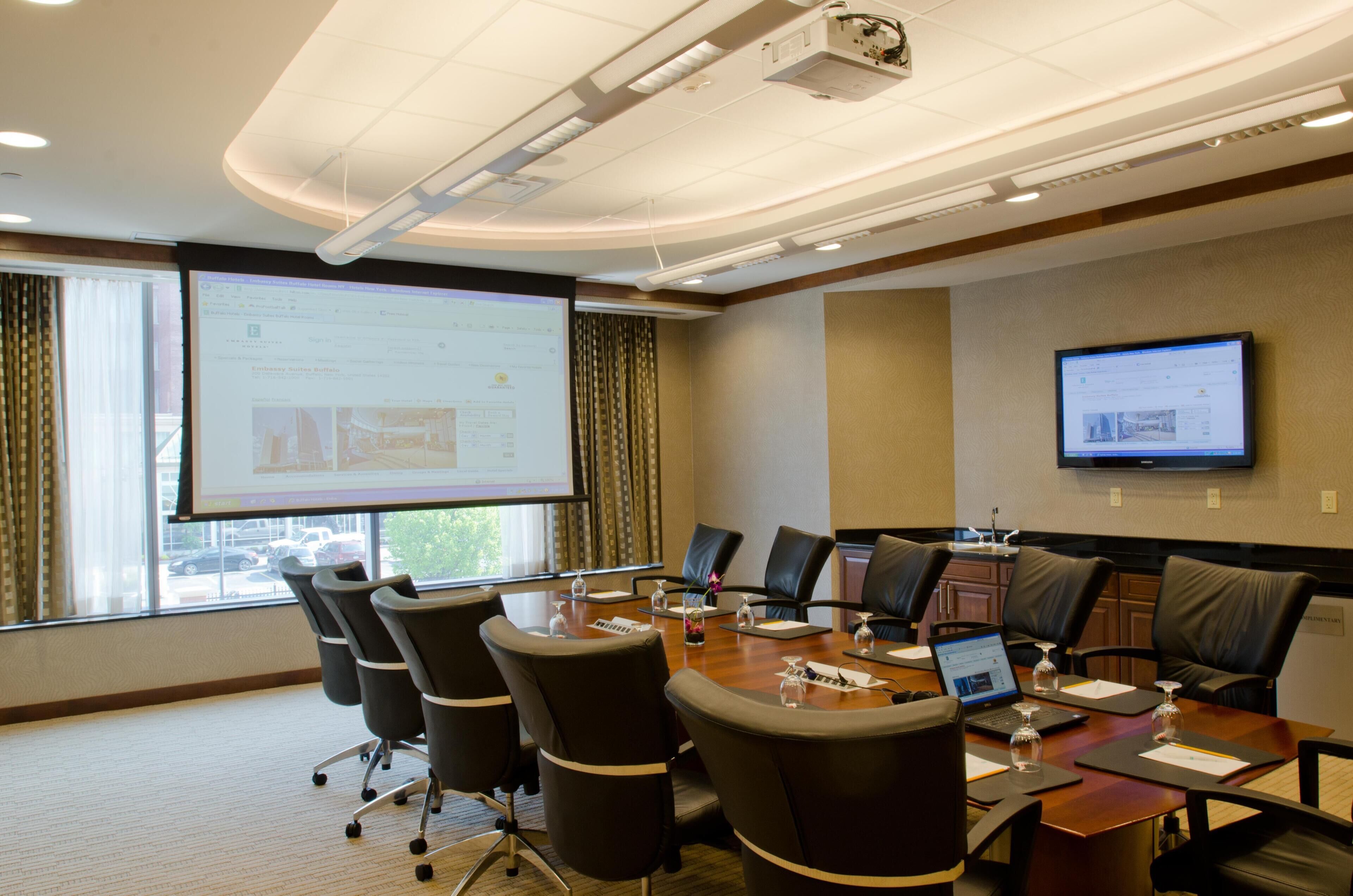 A conference room with a large screen, multiple chairs, a table with laptops, and a television on the wall.