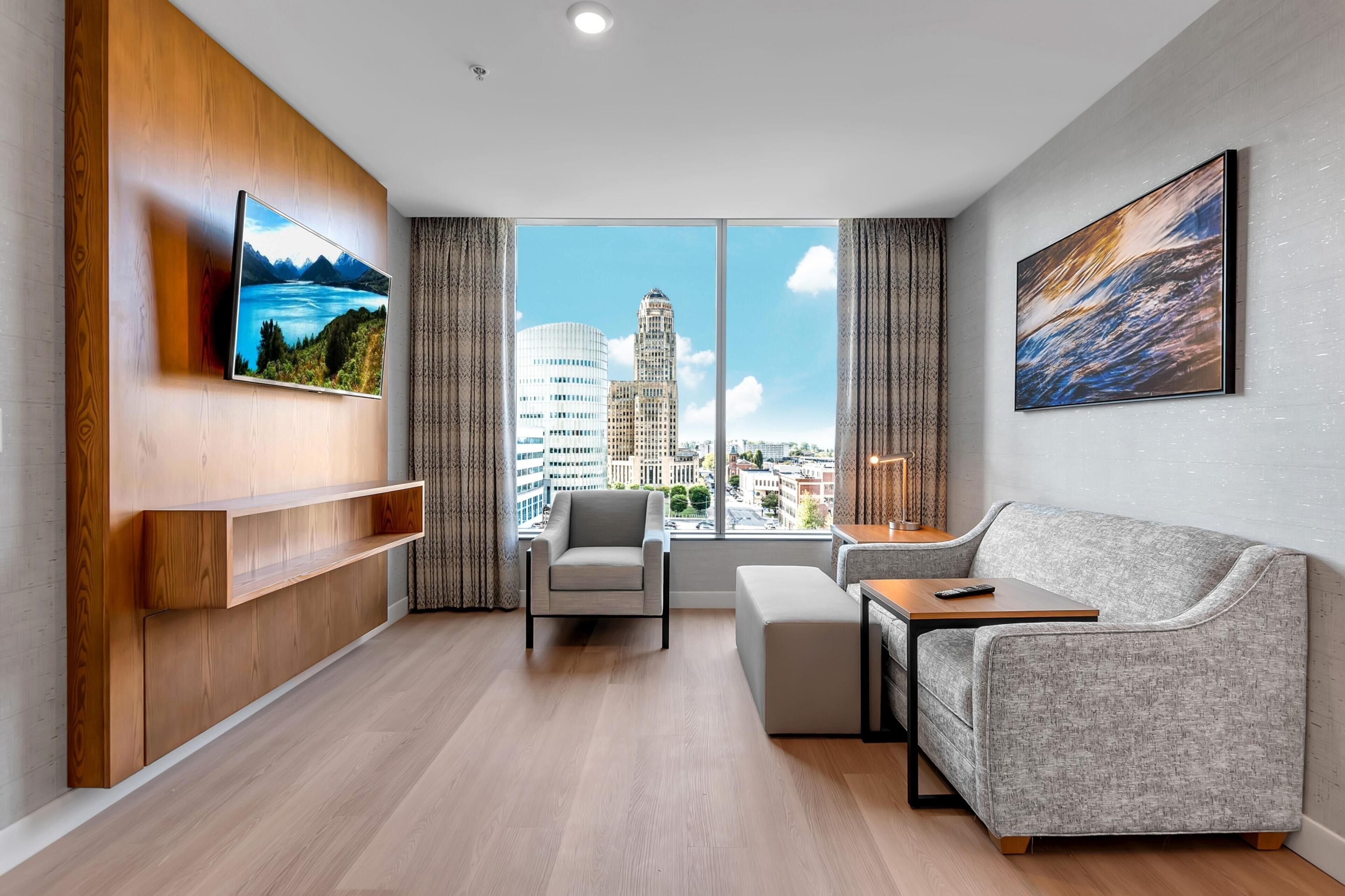 Una sala de estar moderna con un sofá, un sillón y una mesa de centro, un televisor montado en un panel de madera y un ventanal con vistas a la ciudad.