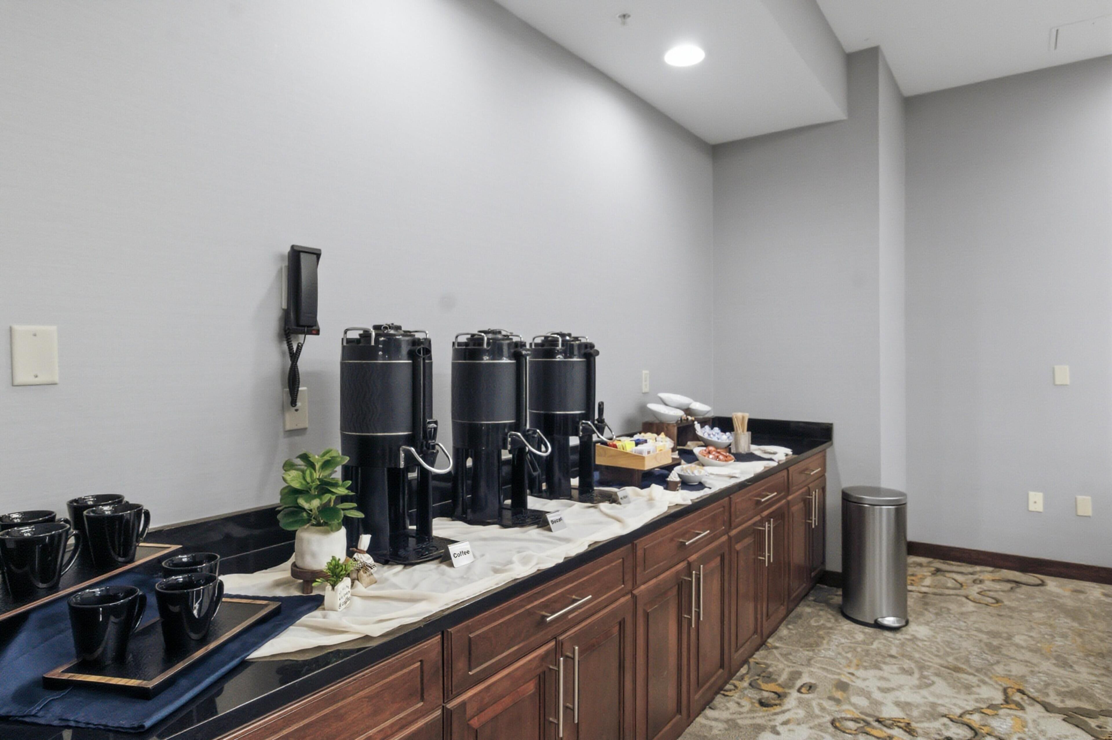 A hotel kitchen with coffee dispensers, a trash can, and a countertop with black cups.