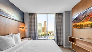 Habitación de hotel con una cama grande, televisor montado en la pared, vista a la ciudad desde la ventana y un estante de madera.