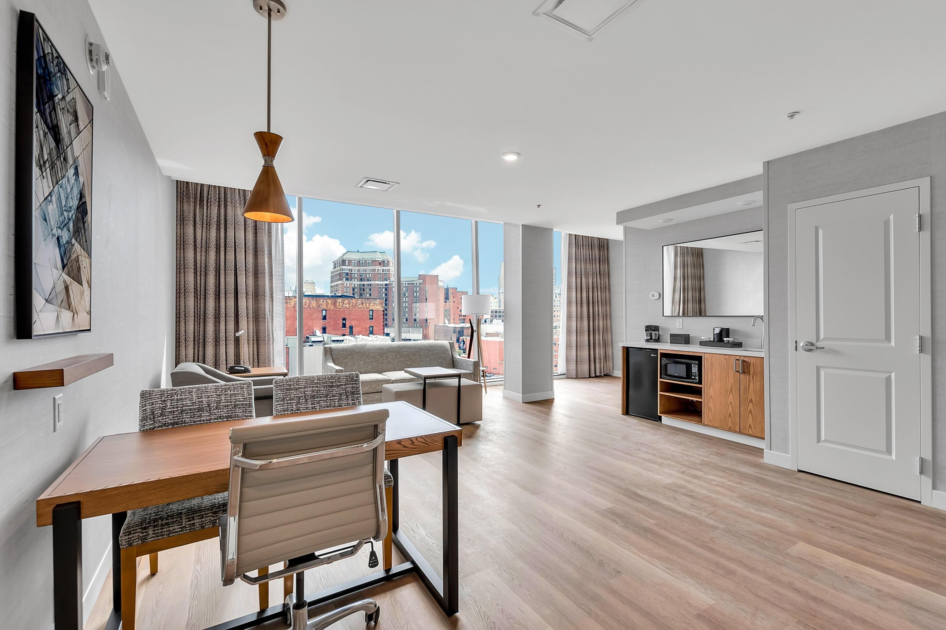 A modern hotel room with a dining area, kitchenette, and large windows offering city views.