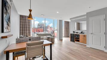 A modern hotel room with a dining area, kitchenette, and large windows offering city views.