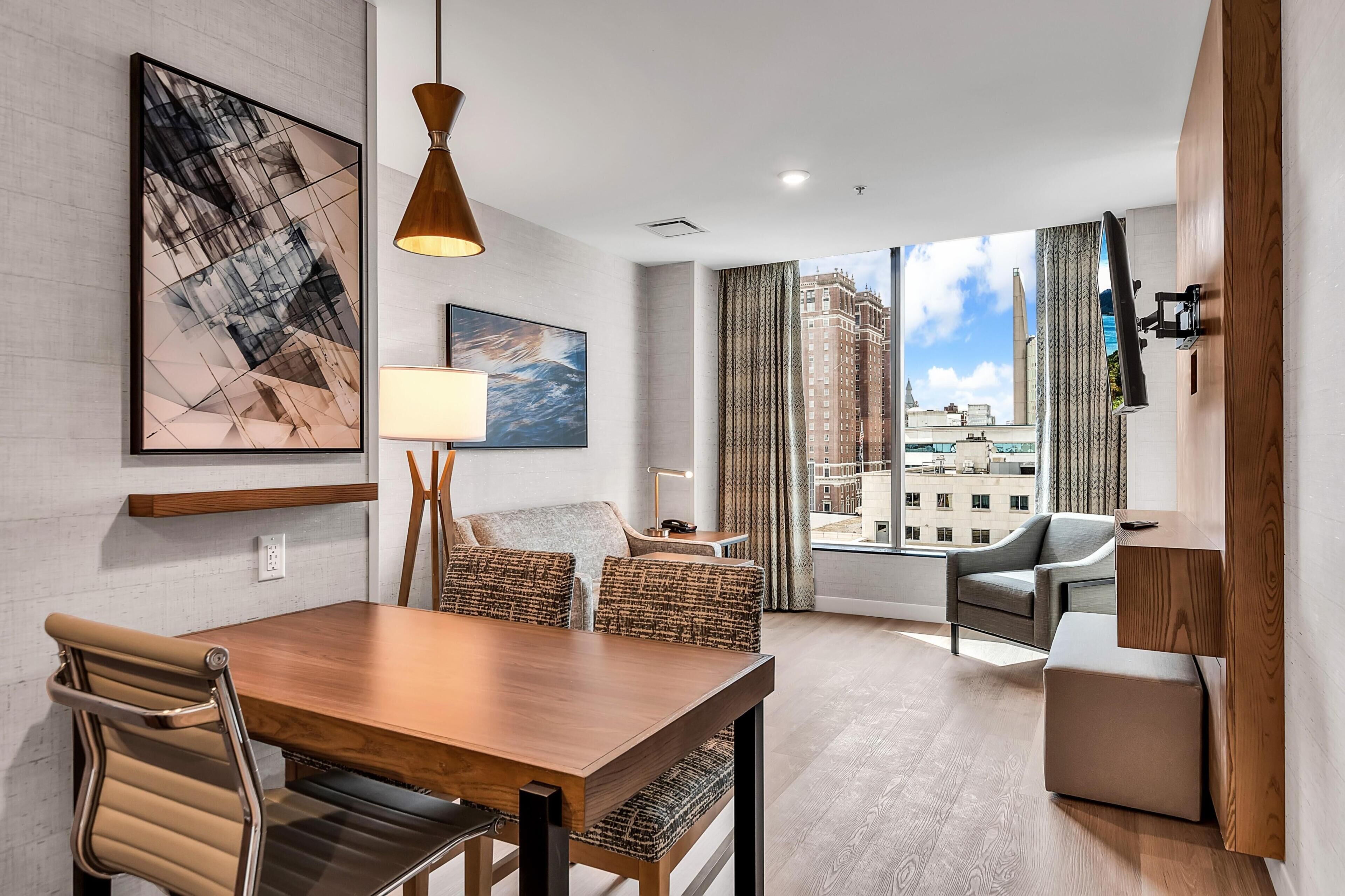 A modern hotel room with a dining area, a sofa, a TV mounted on the wall, and a large window offering a city view.