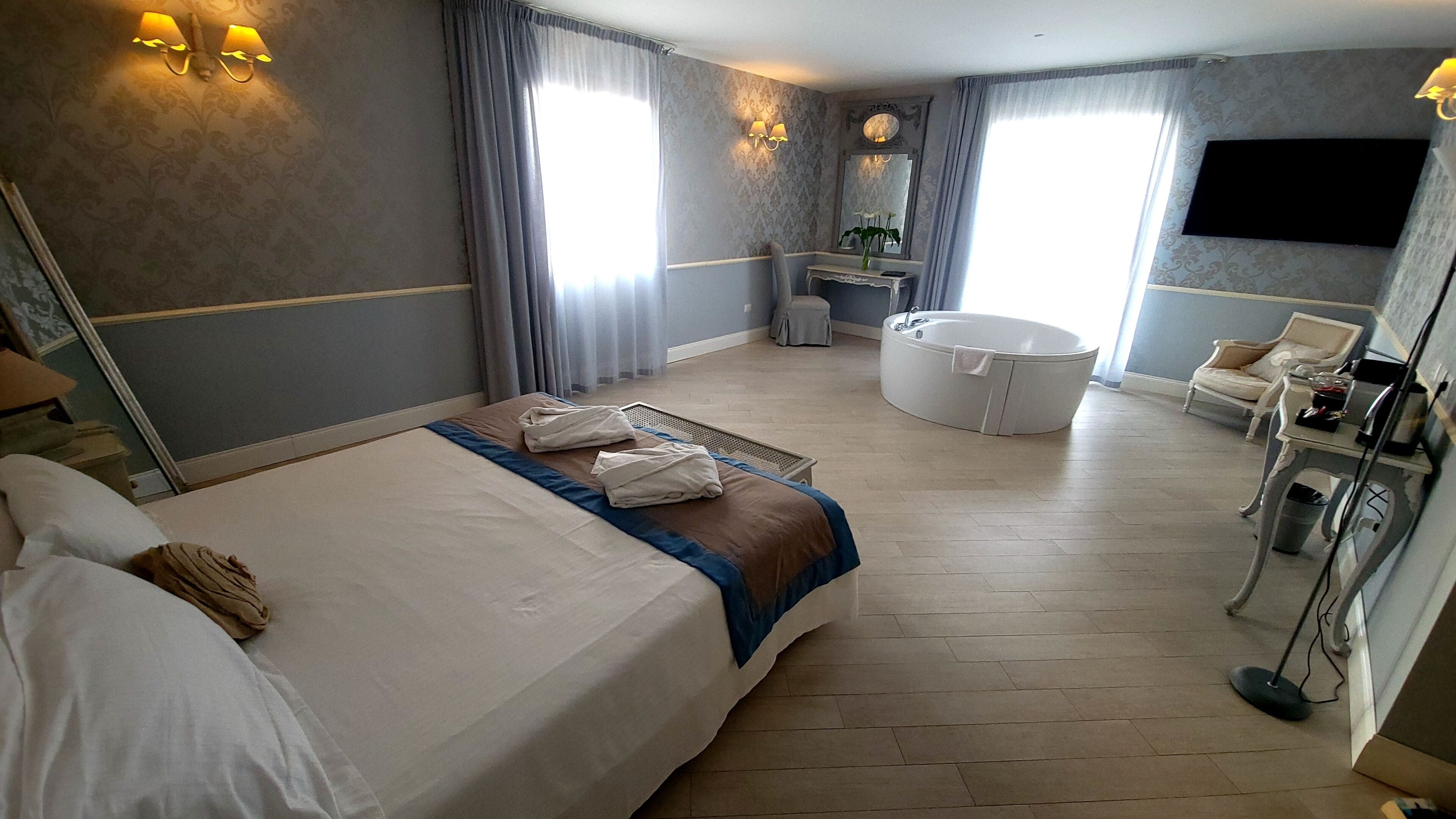 Quarto de hotel com uma cama grande, área de estar com uma poltrona, escrivaninha, banheira e televisão na parede.