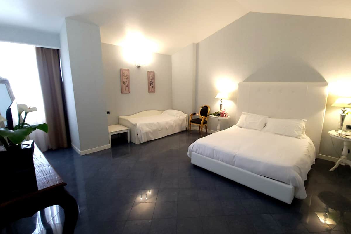 Ein Hotelzimmer mit einem großen Bett, einem kleineren Bett, einem Schreibtisch und einem Sessel. Zwei Wandlampen und ein Beistelltisch mit Lampe gehören ebenfalls zur Ausstattung.