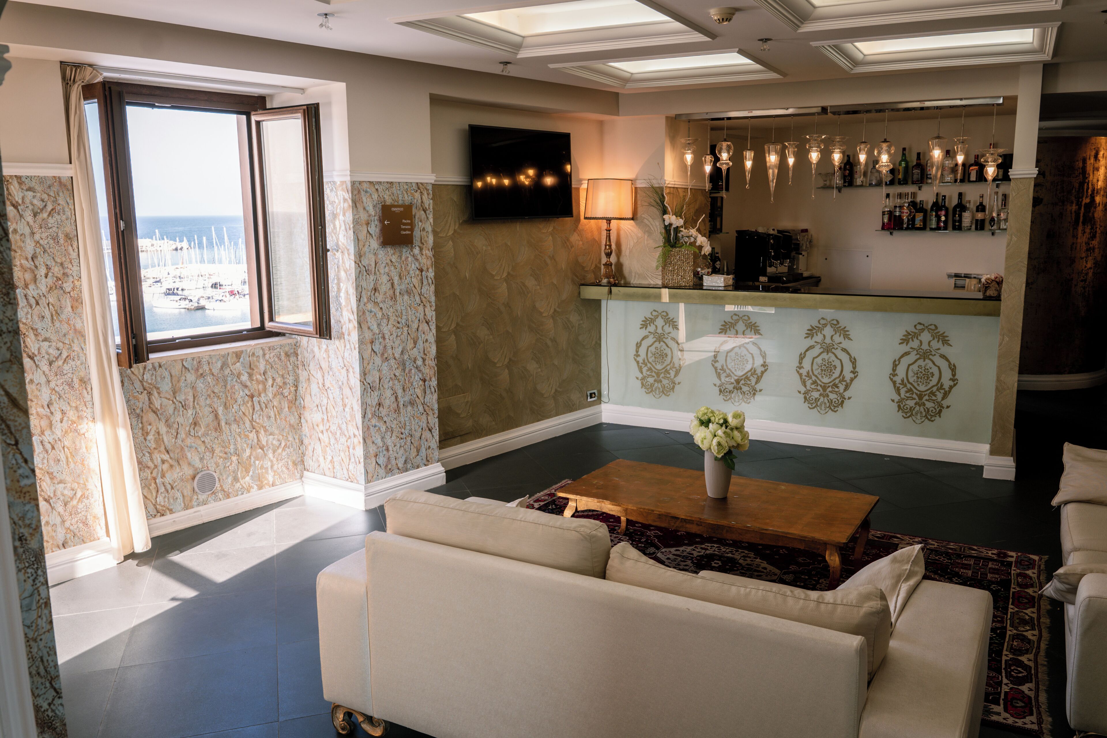 A hotel lobby with a sofa, a coffee table, a TV, a bar area, and a view of the sea through a window.