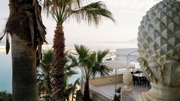 A terrace with palm trees, a stone sculpture, and a view of the sea.