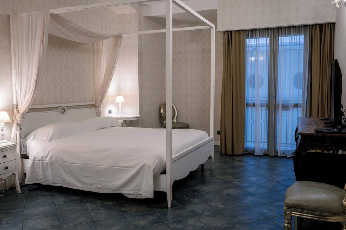 Ein Bett mit Baldachin, ein Nachttisch, ein Beistelltisch, ein Stuhl und ein Blick ins Freie durch ein Fenster.