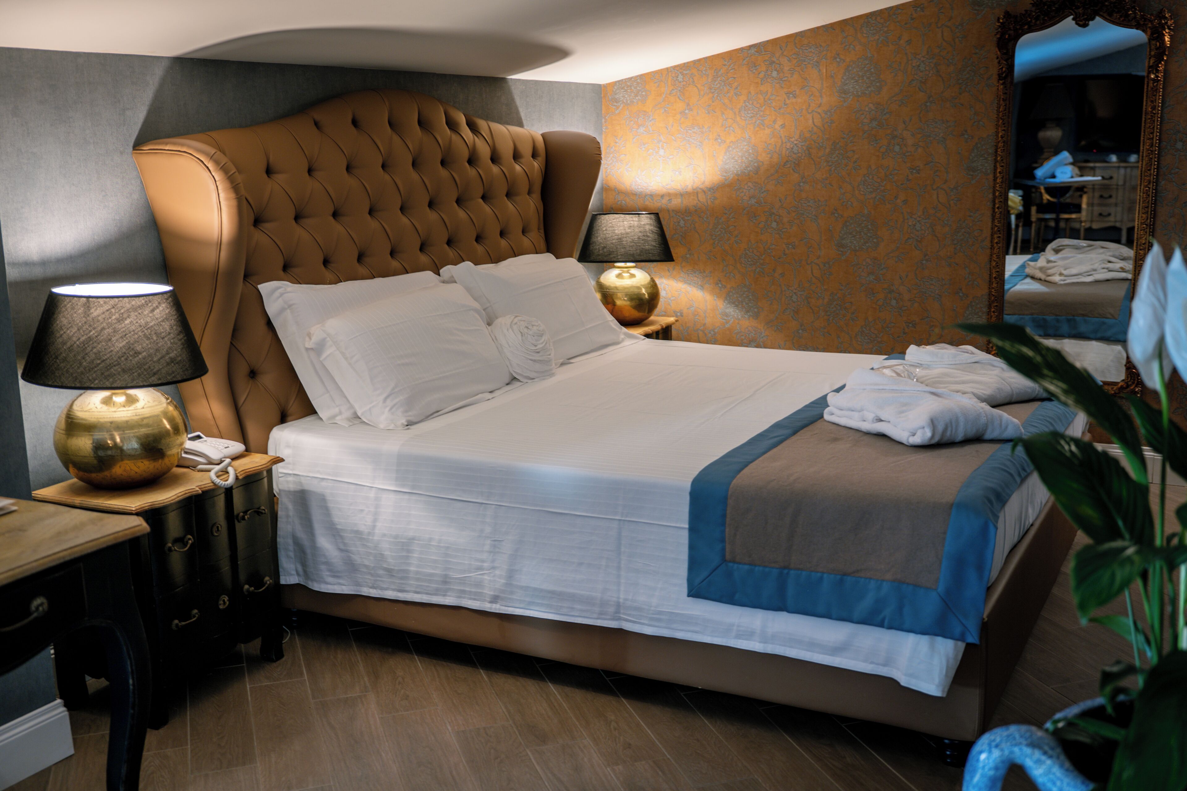 Una habitación de hotel con un cabecero capitonado, una cama grande con ropa blanca, dos lámparas de noche, una mesita de madera y un espejo.