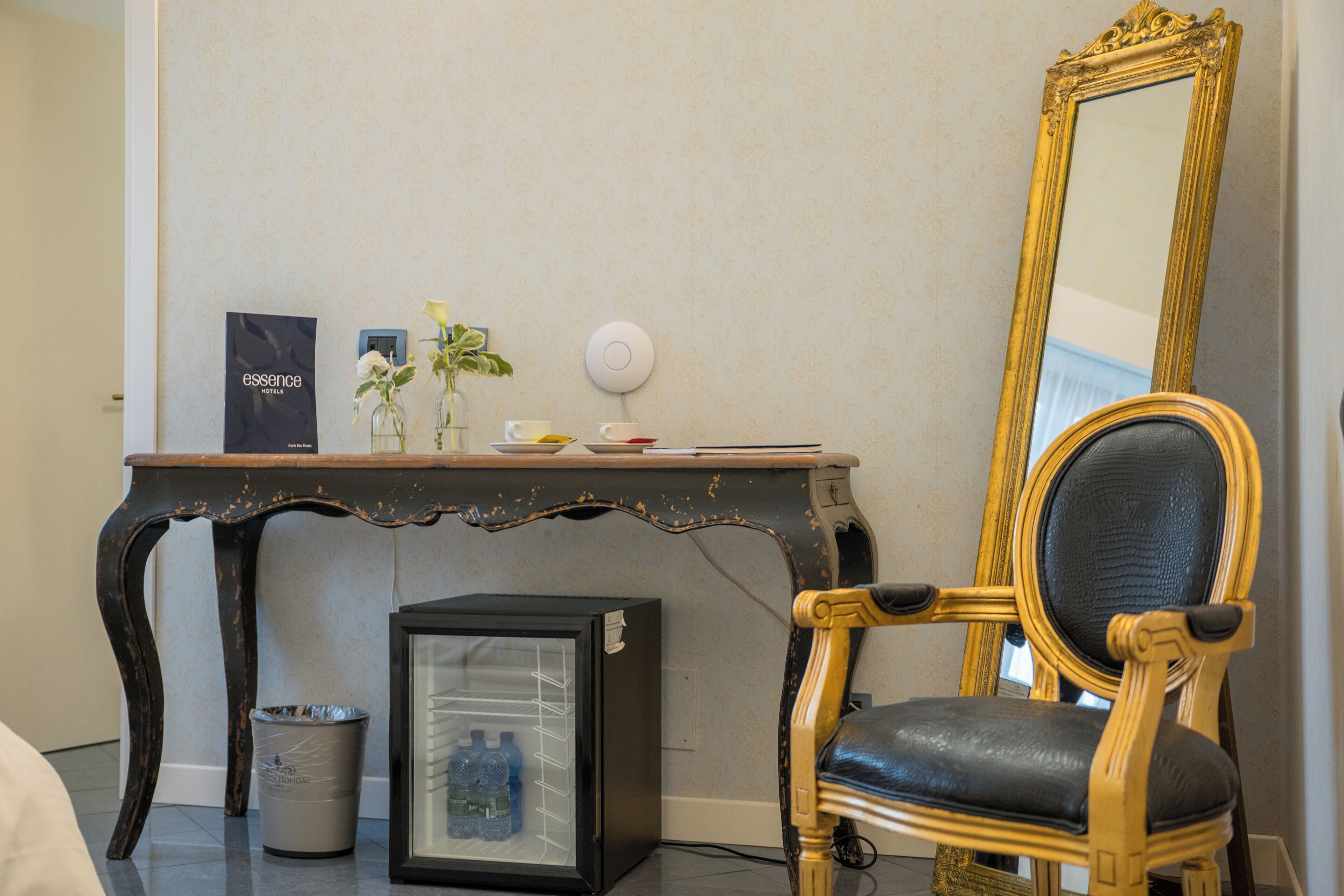 Una habitación con una silla de estilo antiguo, una mesita con un cuadro enmarcado, un cubo de basura y un refrigerador compacto.