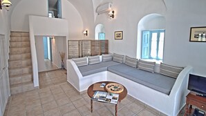 A spacious living area with a large sectional sofa, a small round table, and a staircase leading to an upper level. - Aethrio Sunset Village - Oia (Santorini)