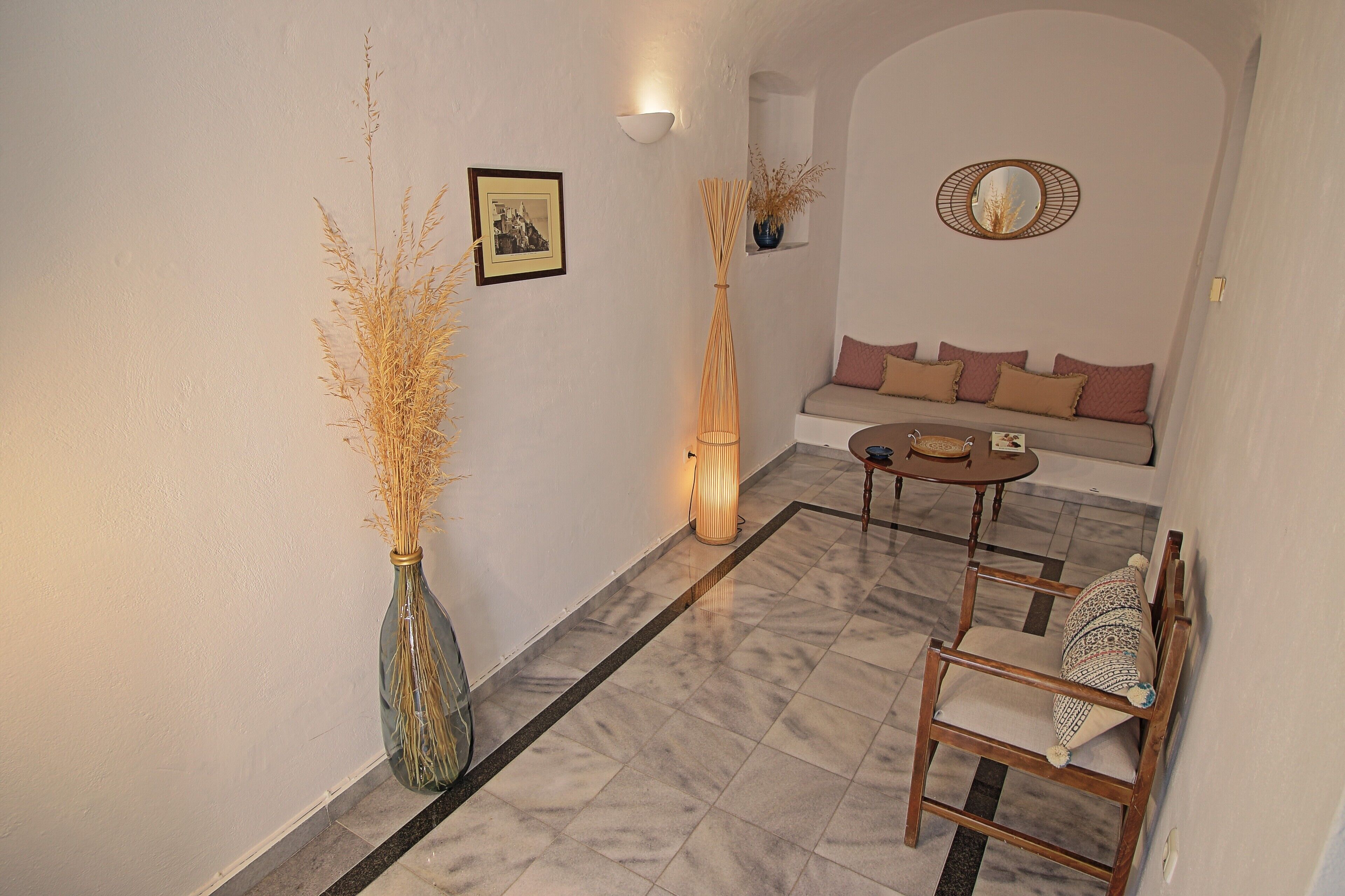 A cozy living area with a sofa, a round coffee table, and a wooden chair. There is a vase with dried plants on the floor and a framed picture on the wall.