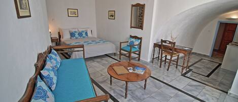 A room with a bed, a sofa, a small table, and a chair. There are framed pictures on the walls and a mirror.