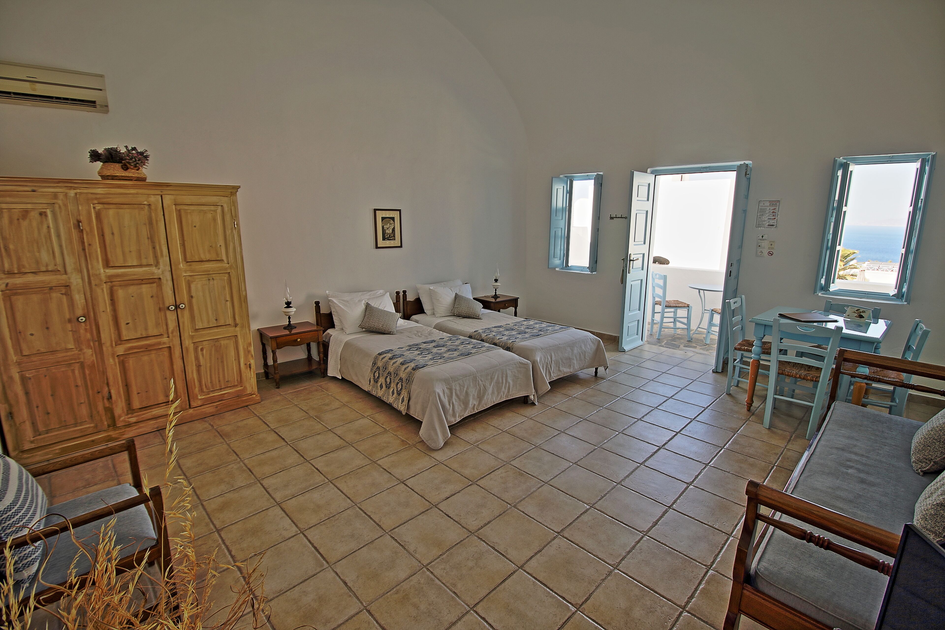 Une chambre avec deux lits, une armoire en bois, un coin repas avec une table et des chaises, et une vue sur la mer par des portes ouvertes.