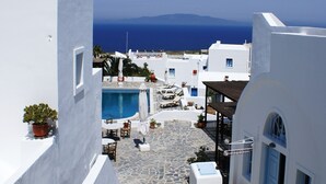 A narrow, white-walled street with a pool and outdoor seating, leading to a view of the sea. - Aethrio Sunset Village - Oia (Santorini)