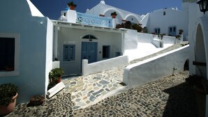 A white building with a blue door and a stone-paved courtyard. - Aethrio Sunset Village - Oia (Santorini)