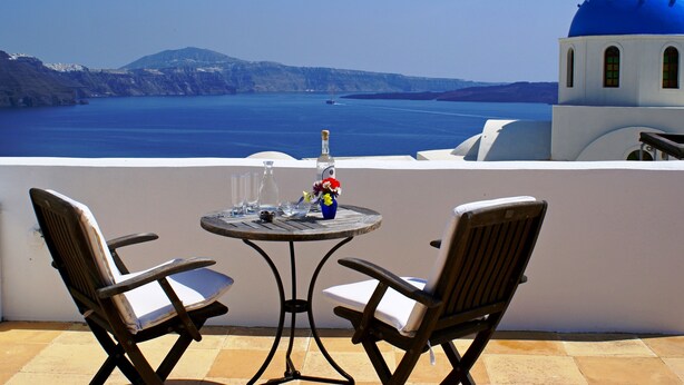 A terrace with two wooden chairs and a table, overlooking a blue sea and a white building with a blue dome. - Aethrio Sunset Village - Oia (Santorini)