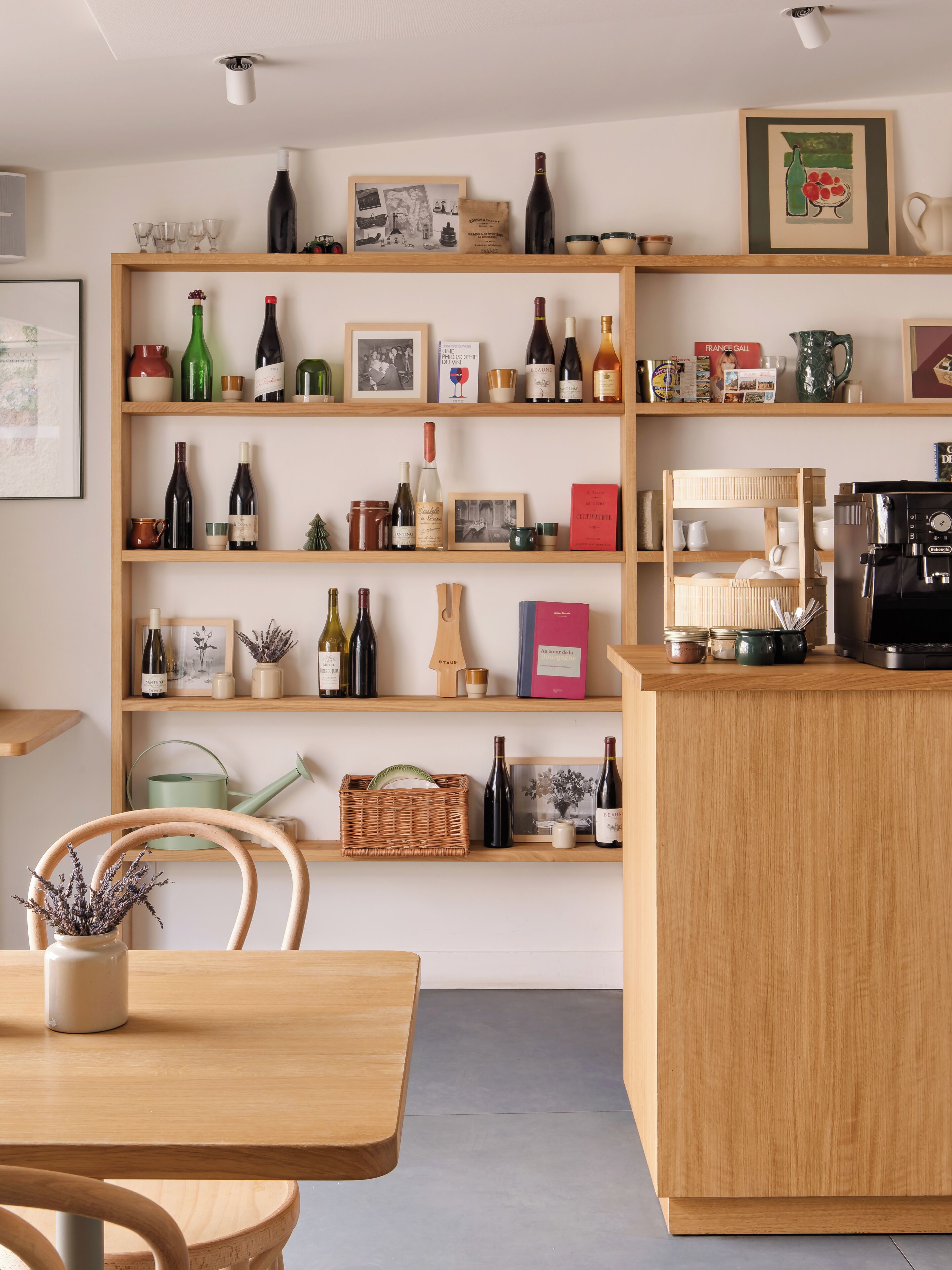 A room with a wooden table, chairs, a coffee machine, and shelves with wine bottles and books.