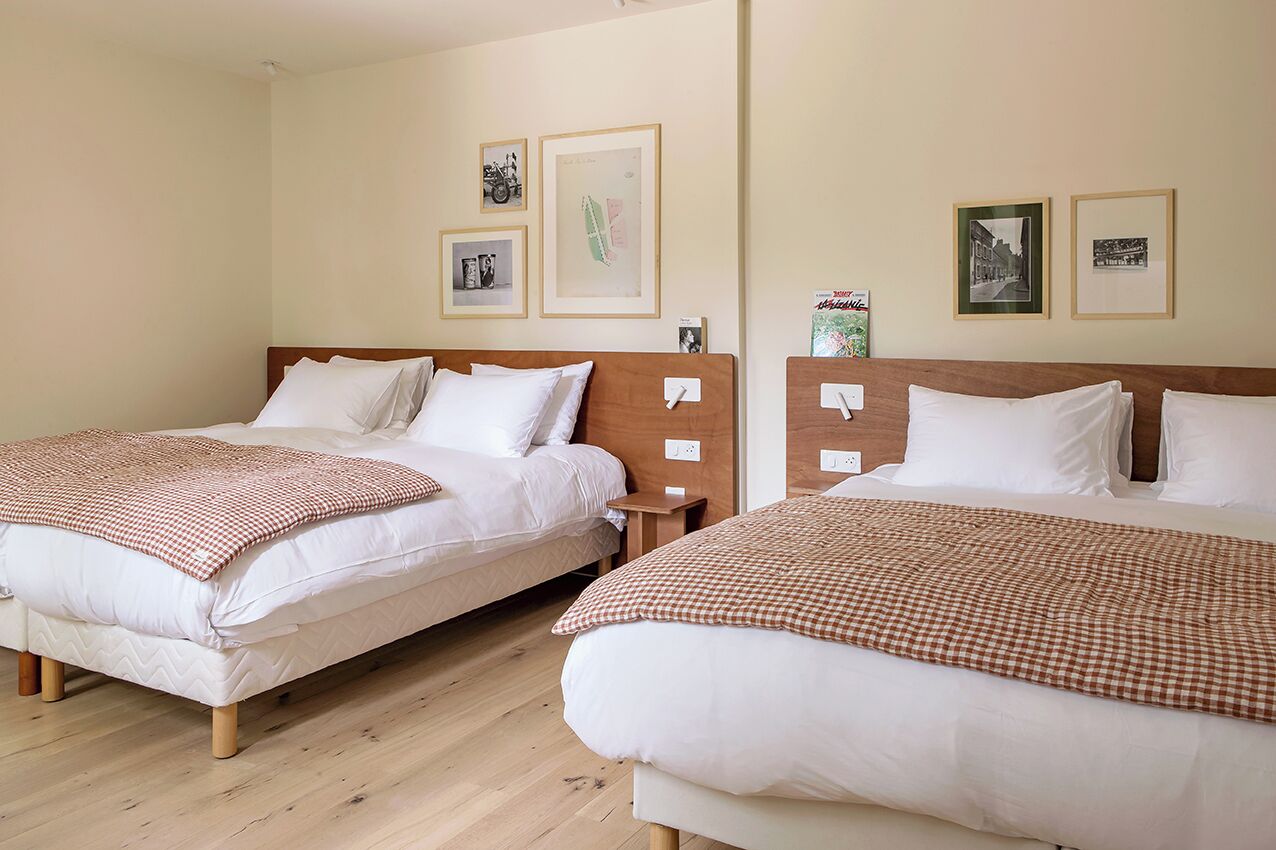 Dos camas individuales con cabeceros de madera, ropa de cama blanca y mantas a cuadros. Hay cuadros enmarcados en la pared y una mesita de noche al lado de cada cama.