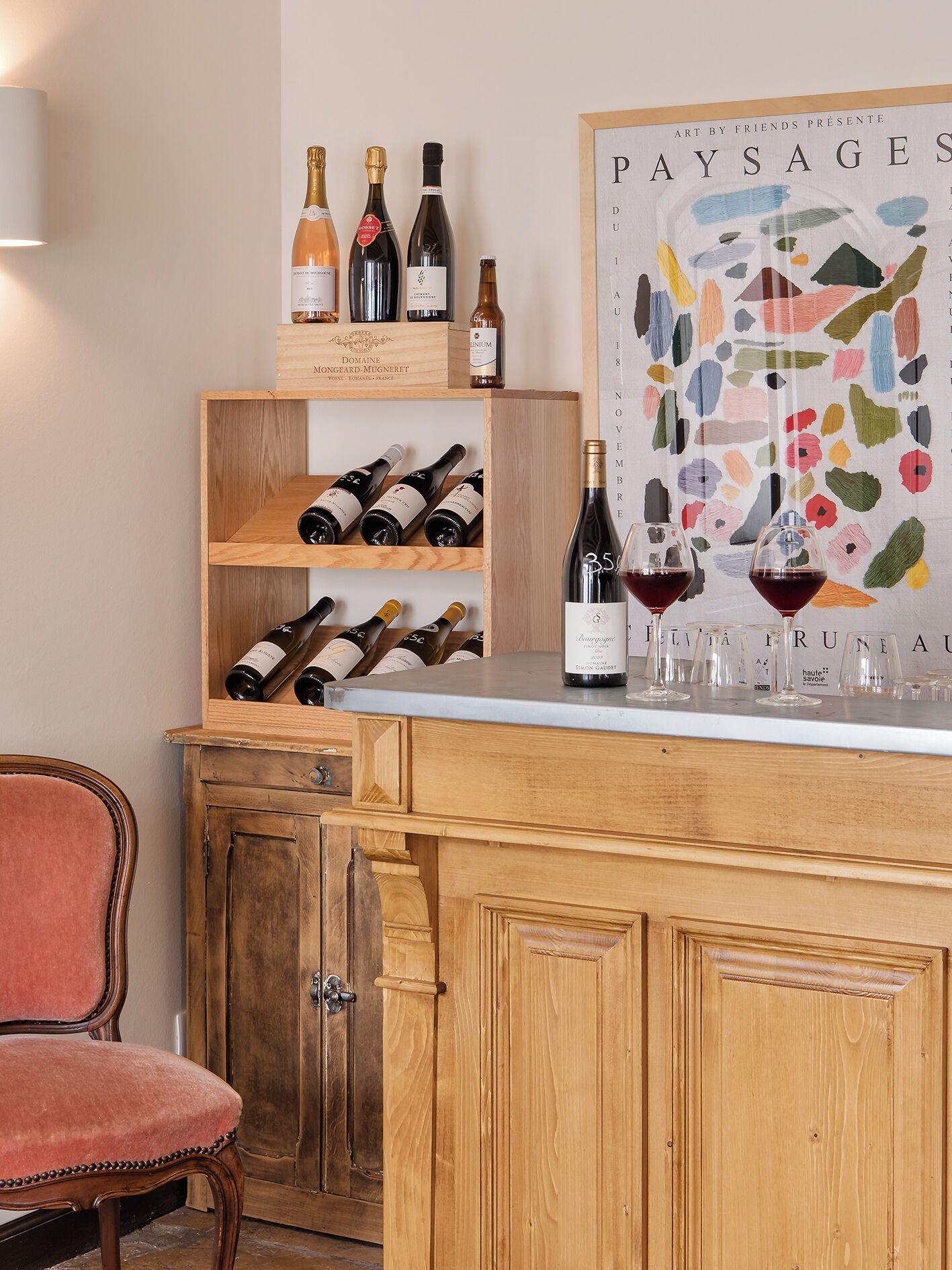 A wooden bar area with wine bottles and glasses, a chair, and a colorful abstract painting.