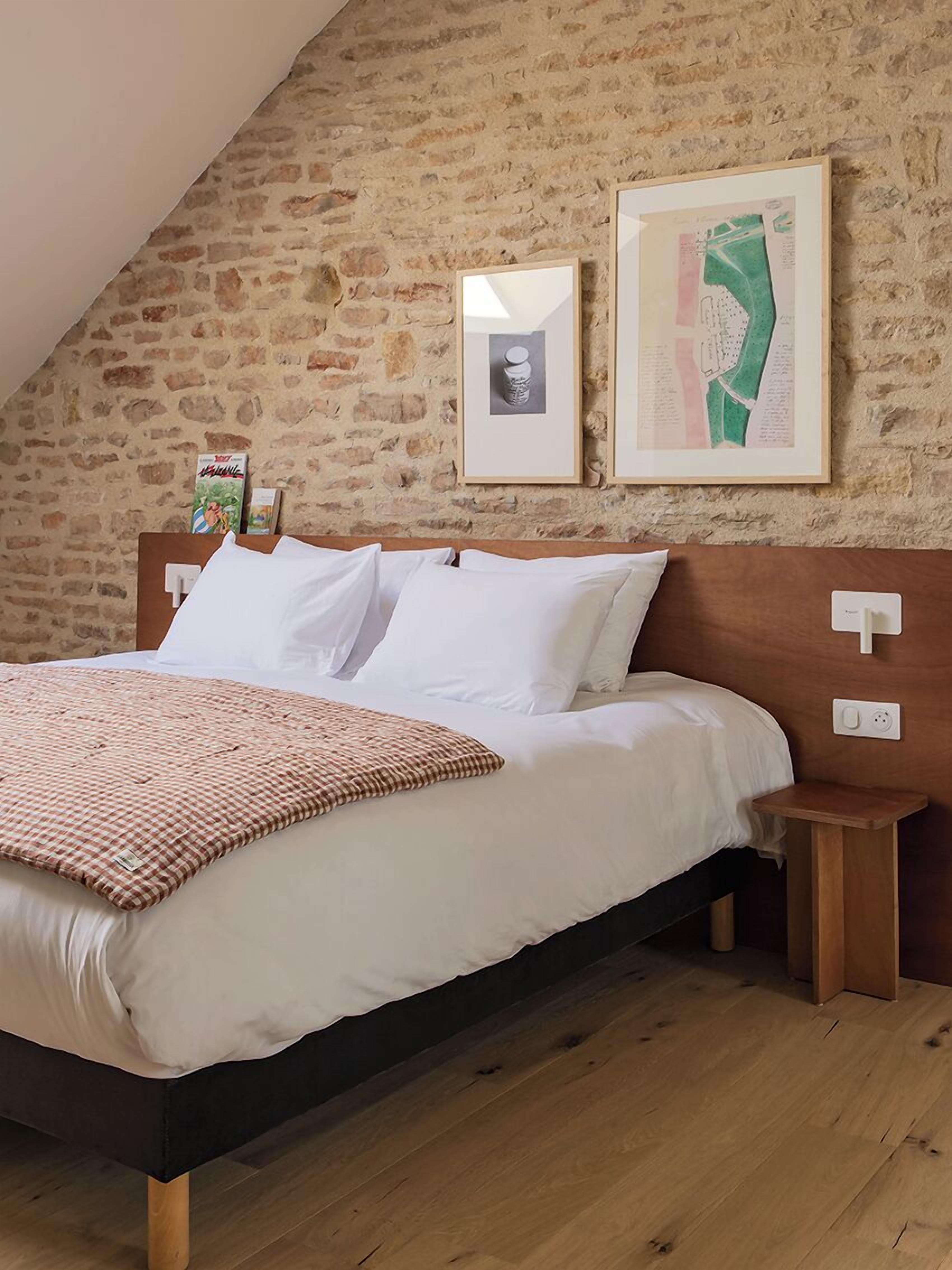 A bedroom with a stone wall, a bed with white linens, a checkered throw blanket, and two framed pictures on the wall.
