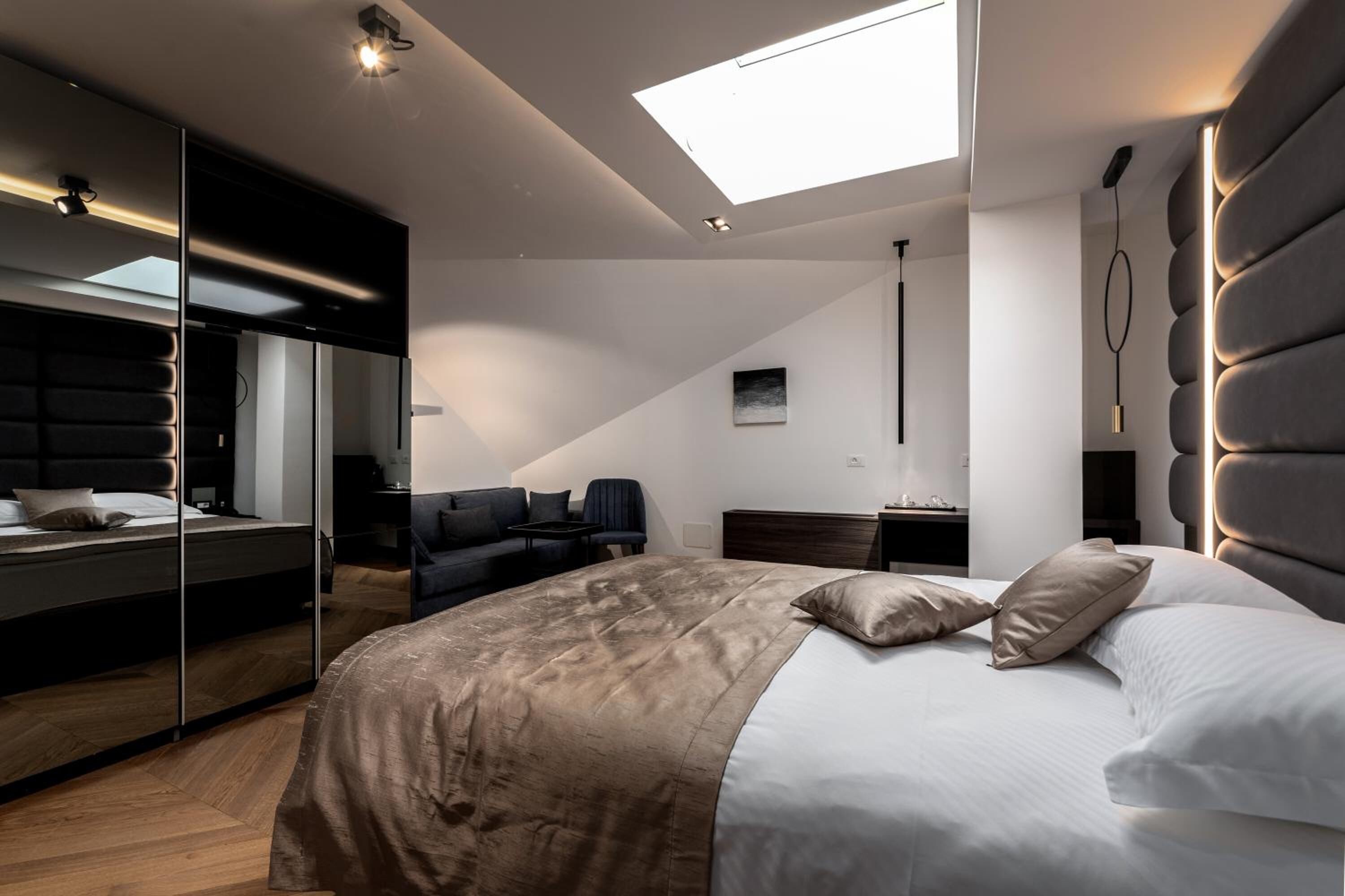 Un dormitorio moderno con una cama grande, un tragaluz, un cabecero oscuro y un área de descanso con sofá y sillones.