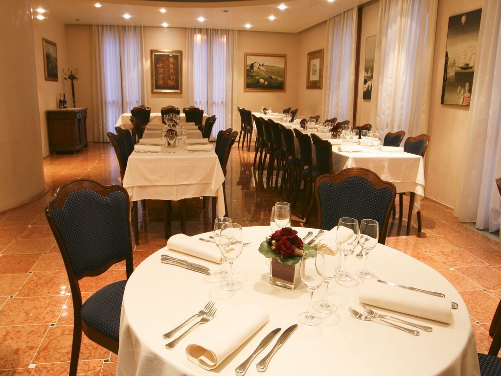 A dining area with round tables set for four, each with white tablecloths, silverware, and wine glasses. The room has a classic design with wooden chairs and a chandelier.