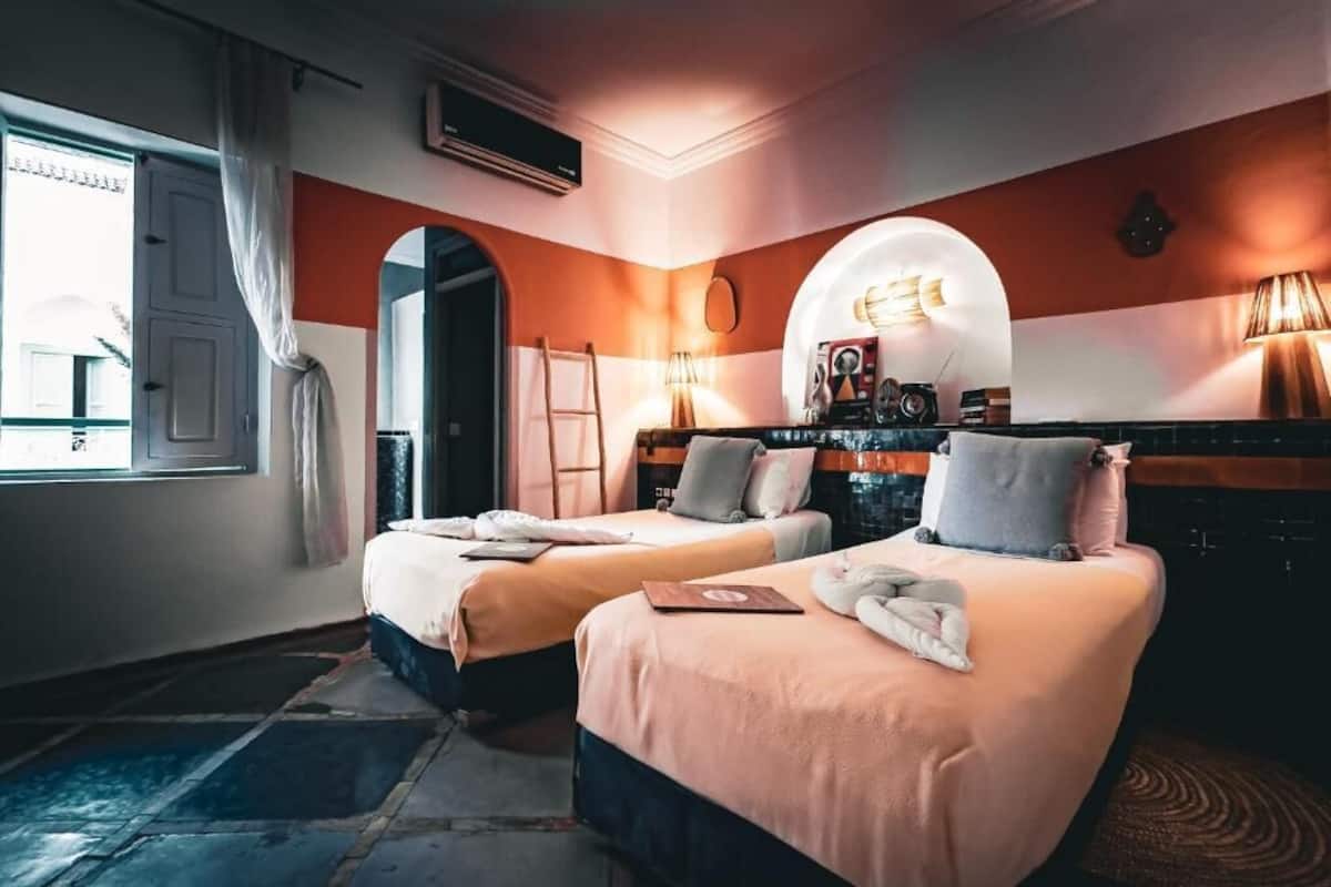 A hotel room with two beds, a window with curtains, a lamp, and a mirror.