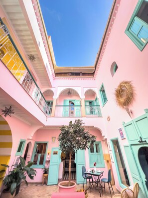 Un patio de varios niveles con paredes rosadas, puertas turquesa y un árbol central.