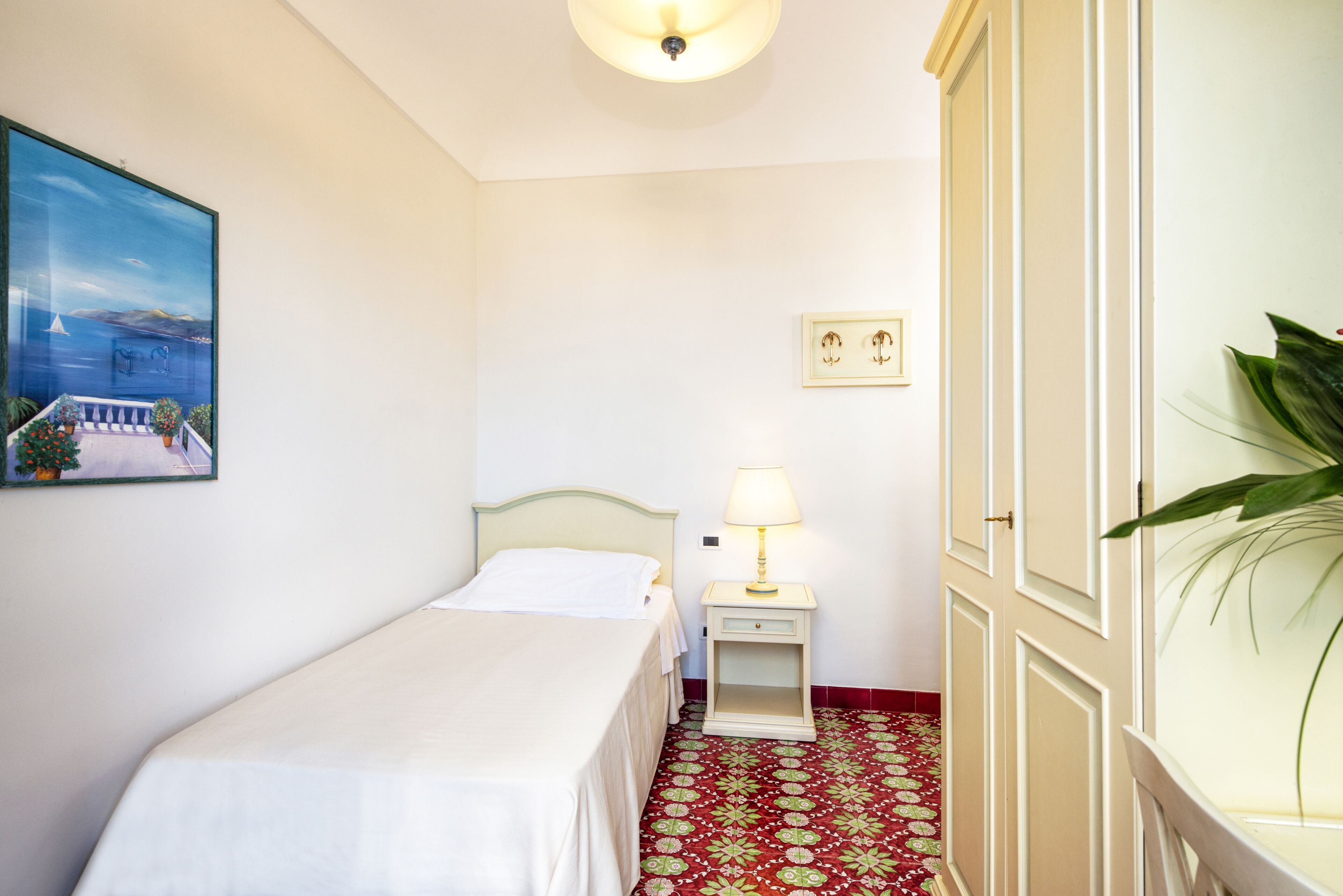 Una habitació d'hotel amb un llit, una petita taula de nit amb una làmpada, un armari, un quadre al mur i una planta en test.
