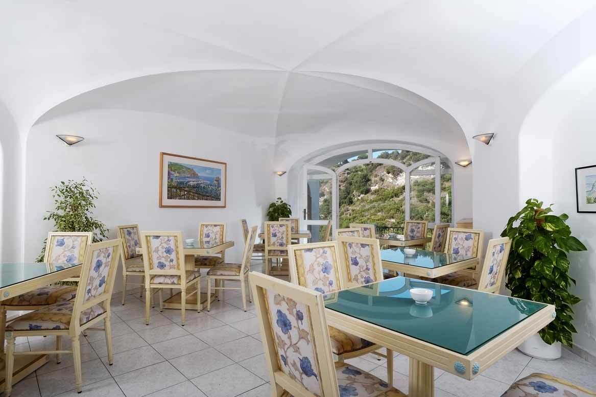 A dining area with a glass table and floral chairs, white walls, and a view of greenery outside.