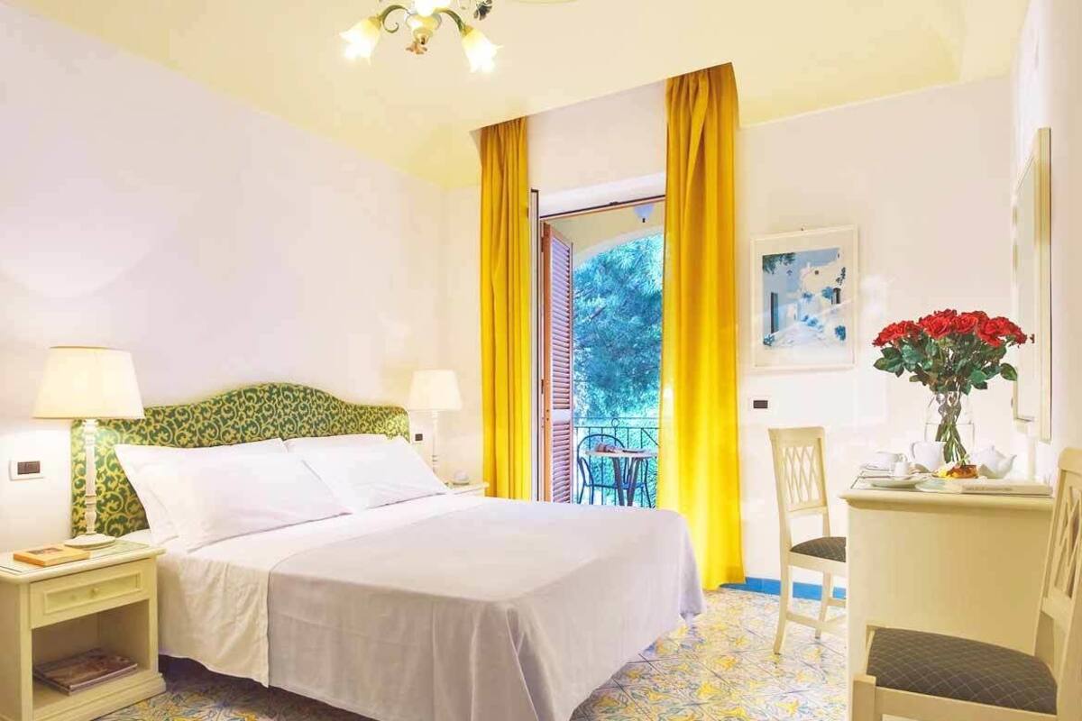 A hotel room with a bed, a nightstand, a chair, a small table with a vase of flowers, and a window with yellow curtains.