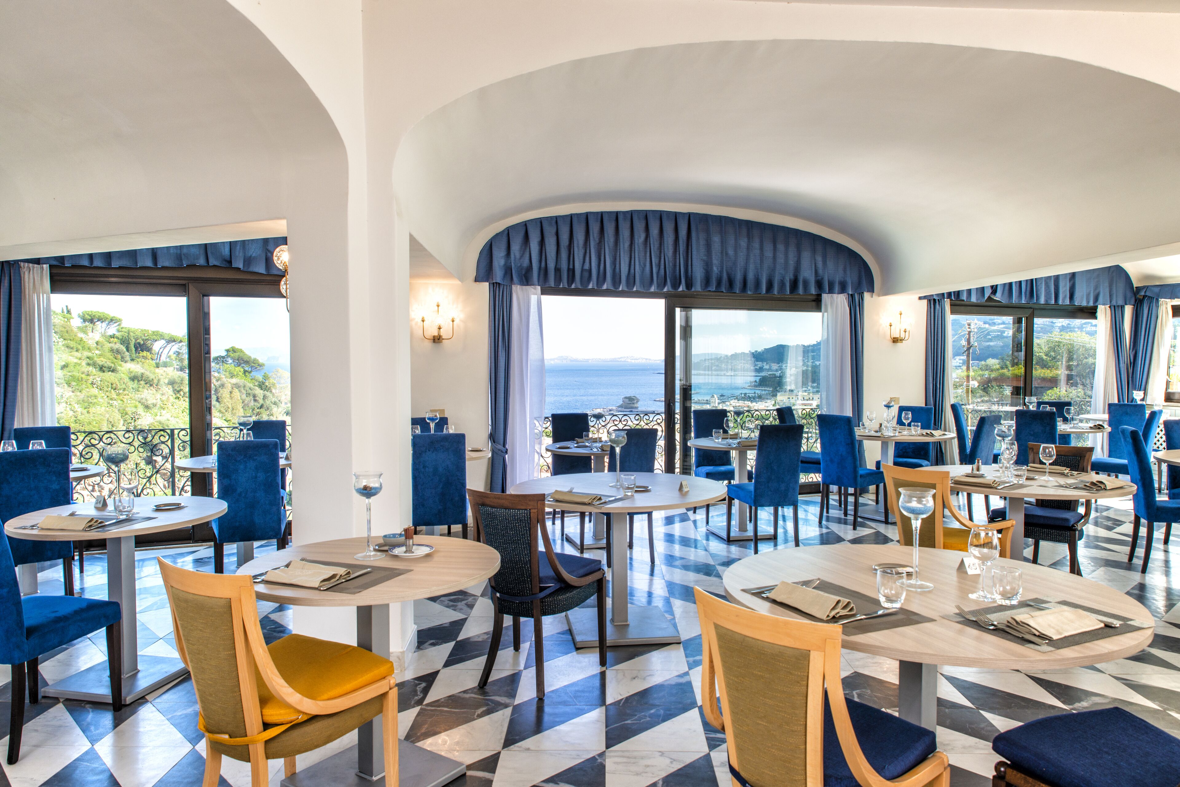 A restaurant with round tables and blue chairs, large windows with curtains, and a checkered floor.