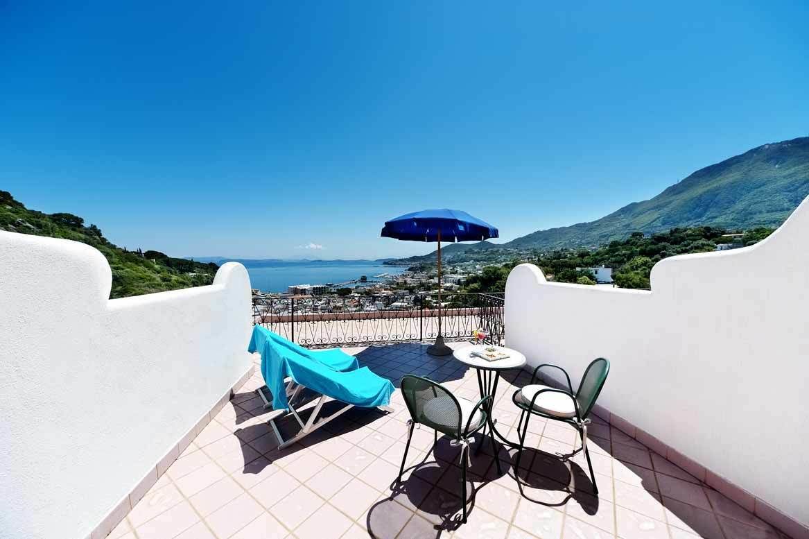 A terrace with a table and chairs, a blue umbrella, and a view of the sea and hills.