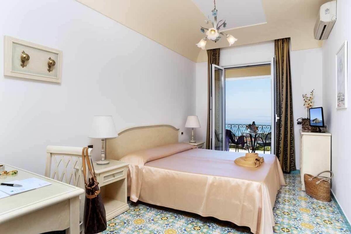 A hotel room with a bed, bedside table, lamp, and a view of the sea through a large window.