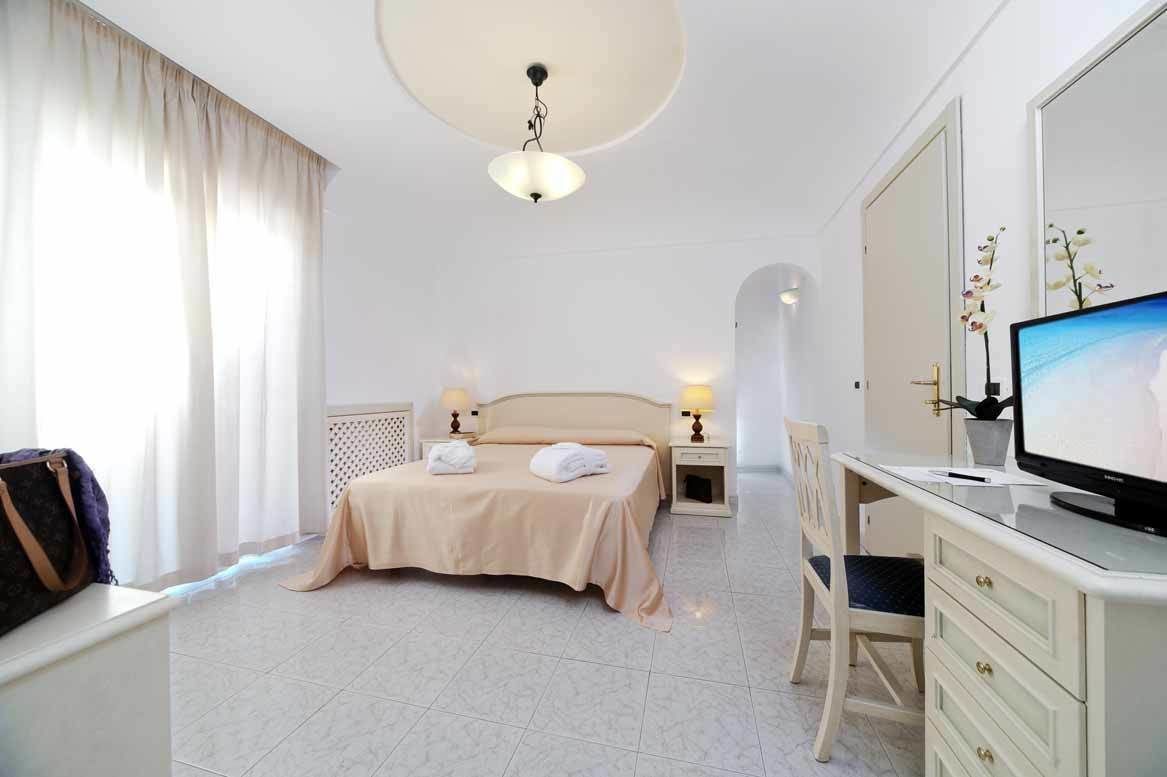 Camera d'albergo con un letto, due lampade ai comodini, una scrivania con un computer, una televisione e una finestra con tende.