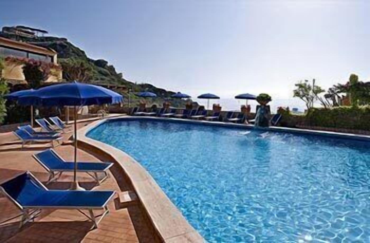 Piscina con sillones de descanso y sombrillas, rodeada de vegetación y un cielo despejado.
