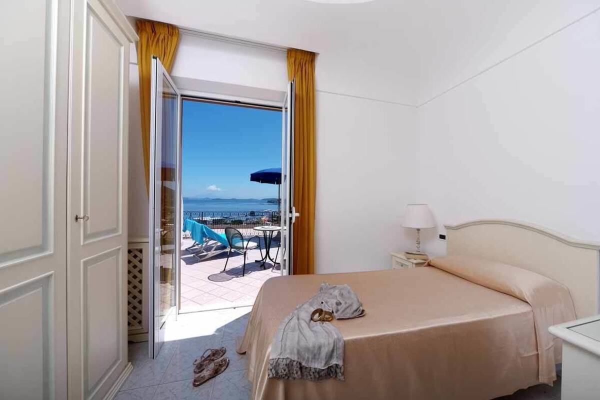A hotel room with a bed, a nightstand, a lamp, and a view of the sea through an open door.