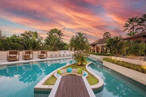 A pool area with a wooden deck and seating, surrounded by palm trees and a sunset sky.