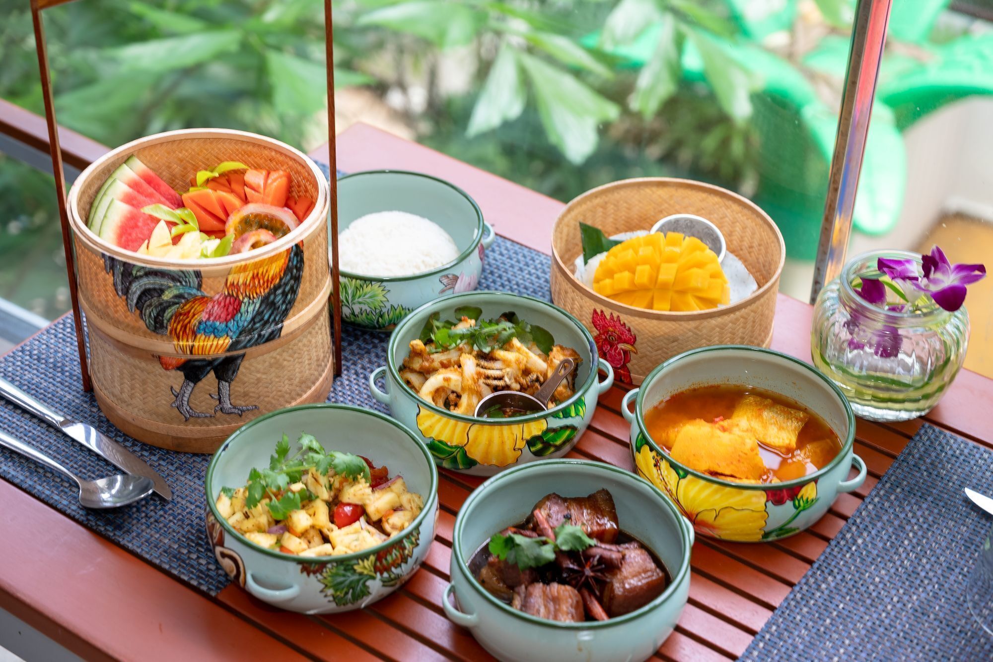 Una mesa con varios platos y recipientes, algunos con diseños coloridos, colocada al aire libre sobre una mesa de madera con vistas a la vegetación.