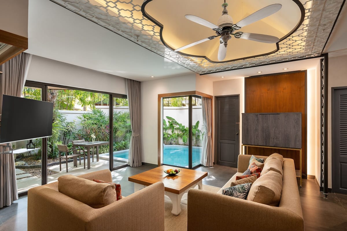 A modern living room with a sofa, coffee table, and TV. There is a pool visible through the glass doors.
