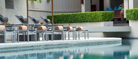 Un espace aménagé au bord de la piscine, avec des chaises longues et des serviettes, un mur en pierre orné d’une plaque commémorative et une haie bien entretenue.