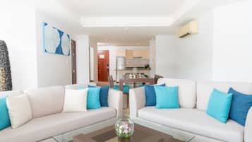Una sala de estar moderna con una mesa de centro de vidrio, dos sofás blancos con cojines azules y una cocina al fondo.