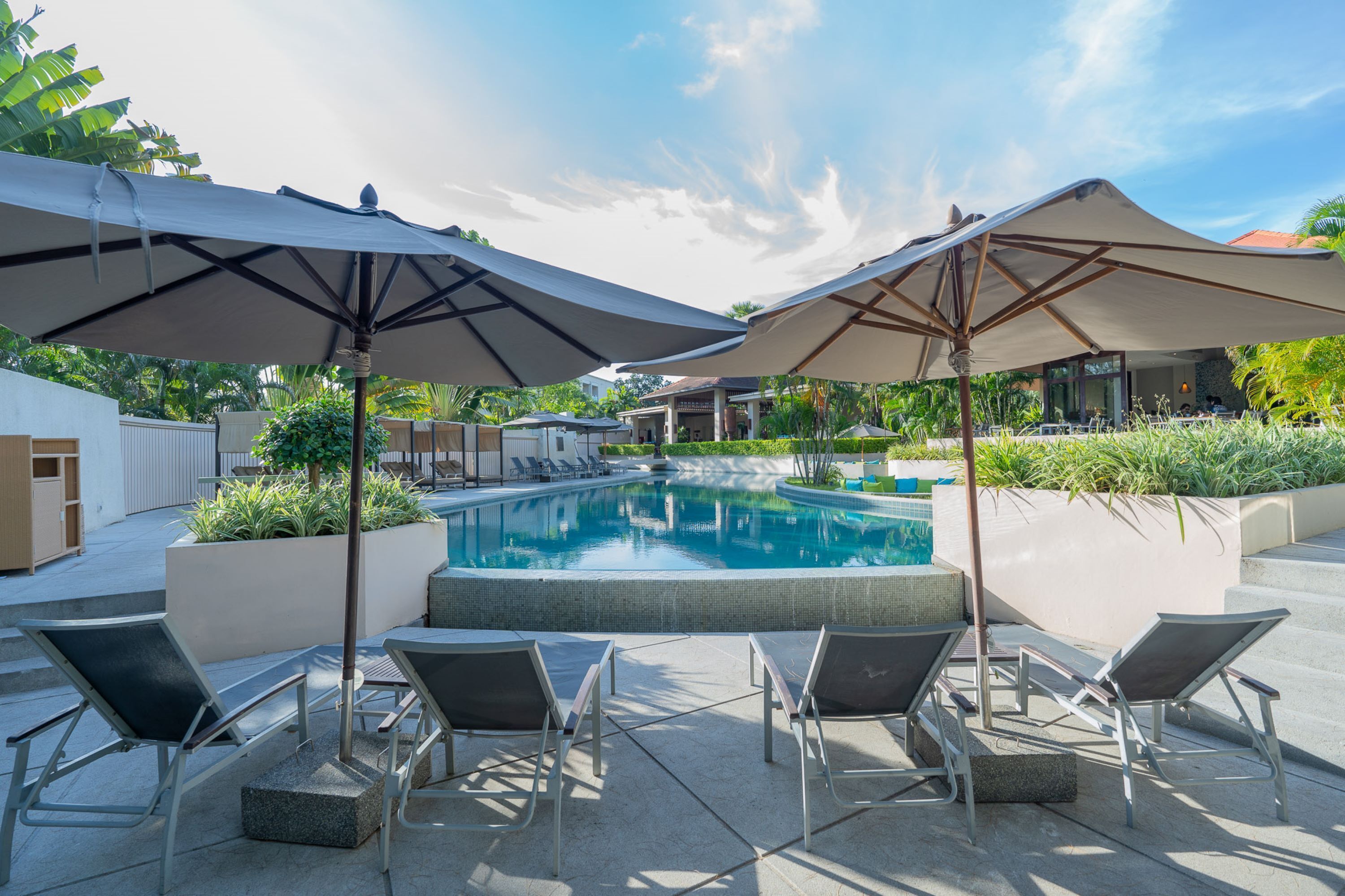 Een zwembadterras met ligstoelen en parasols, omgeven door groen en een gebouw op de achtergrond.