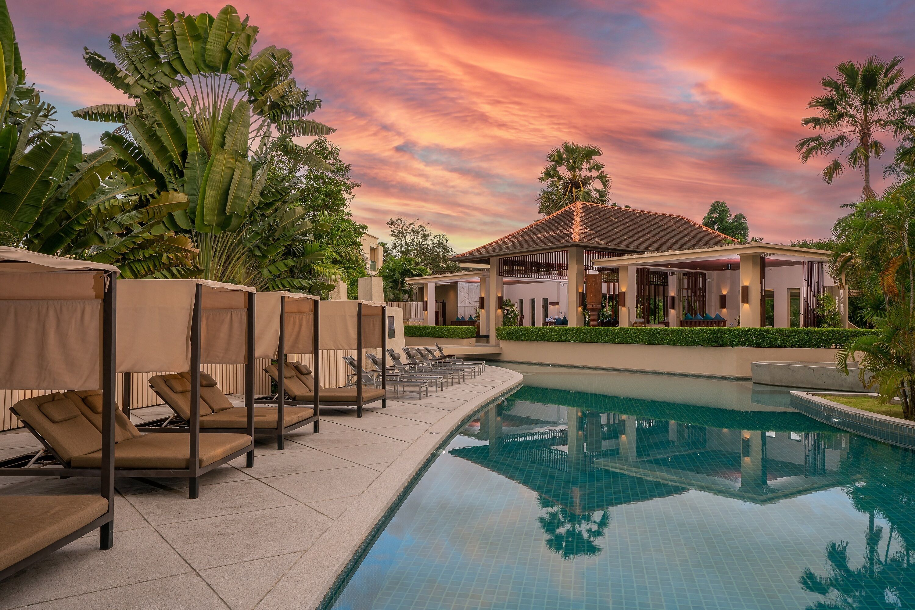 A pool area with lounge chairs and a covered pavilion, surrounded by tropical plants.