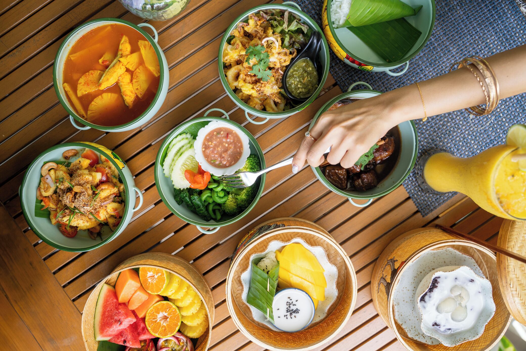 A variety of dishes on a wooden table, including bowls with different foods and a hand reaching for one of the bowls.