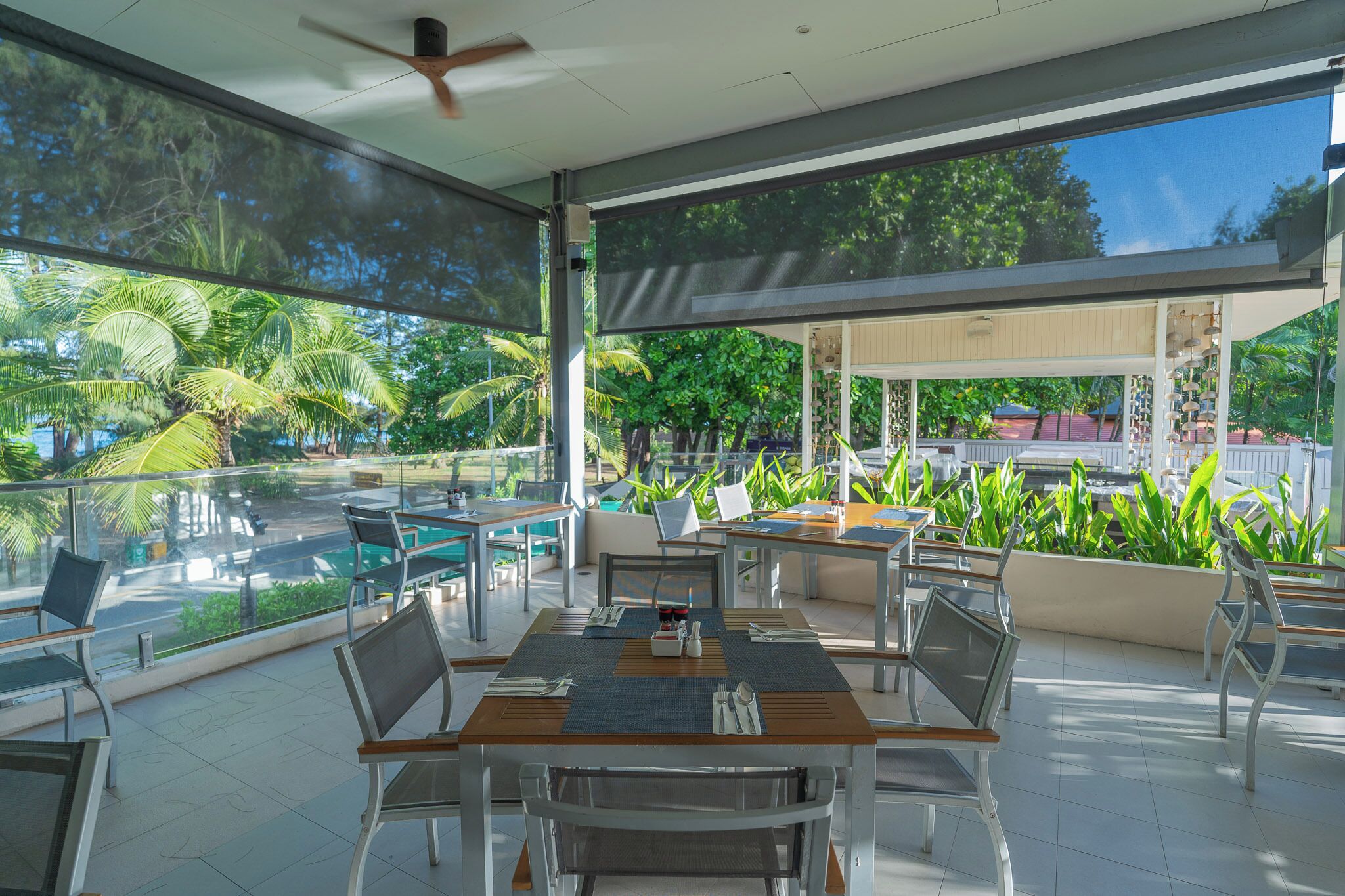 A spacious outdoor dining area with tables and chairs, surrounded by large windows offering views of lush greenery and palm trees.