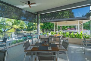 Amplia zona de comedor al aire libre con mesas y sillas, rodeada por grandes ventanales que ofrecen vistas a exuberante vegetación y palmeras.
