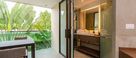 A modern bathroom with a glass shower enclosure, a wooden vanity, and a view of a balcony with large green leaves.